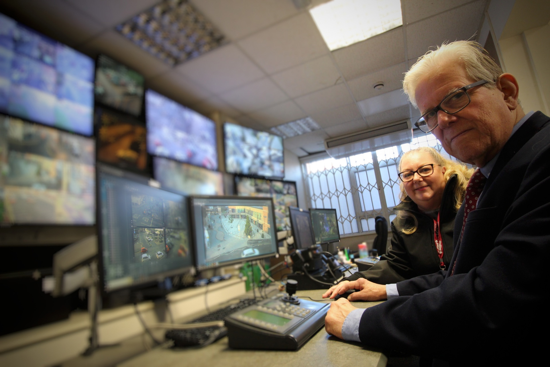 John Clarke MBE in the council CCTV Control Room. Monitors and controlls are on the left hand side with screens blurred out. Cllr John Clarke MBE and Council Officer are sat on the right side looking at the camera.